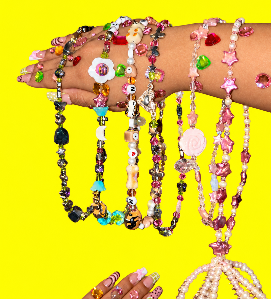 Colorful handmade necklaces with stars, crystals, flowers, shells and playful beads draped over jeweled hands against a vivid yellow background.